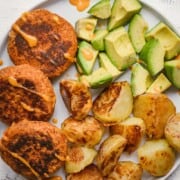 Sun-Dried Tomato Tempeh Burgers served with crispy oven-baked potatoes and avocado chunks.