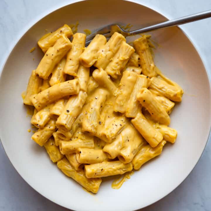 Rigatoni pasta with creamy vegan lemon sauce, topped with lemon zest, black pepper, and crushed red pepper in a blue bowl with a fork.