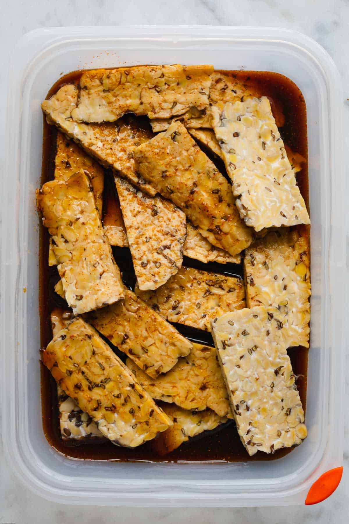 Tempeh cut into rectangles, marinating in soy maple marinade in a Tupperware container.