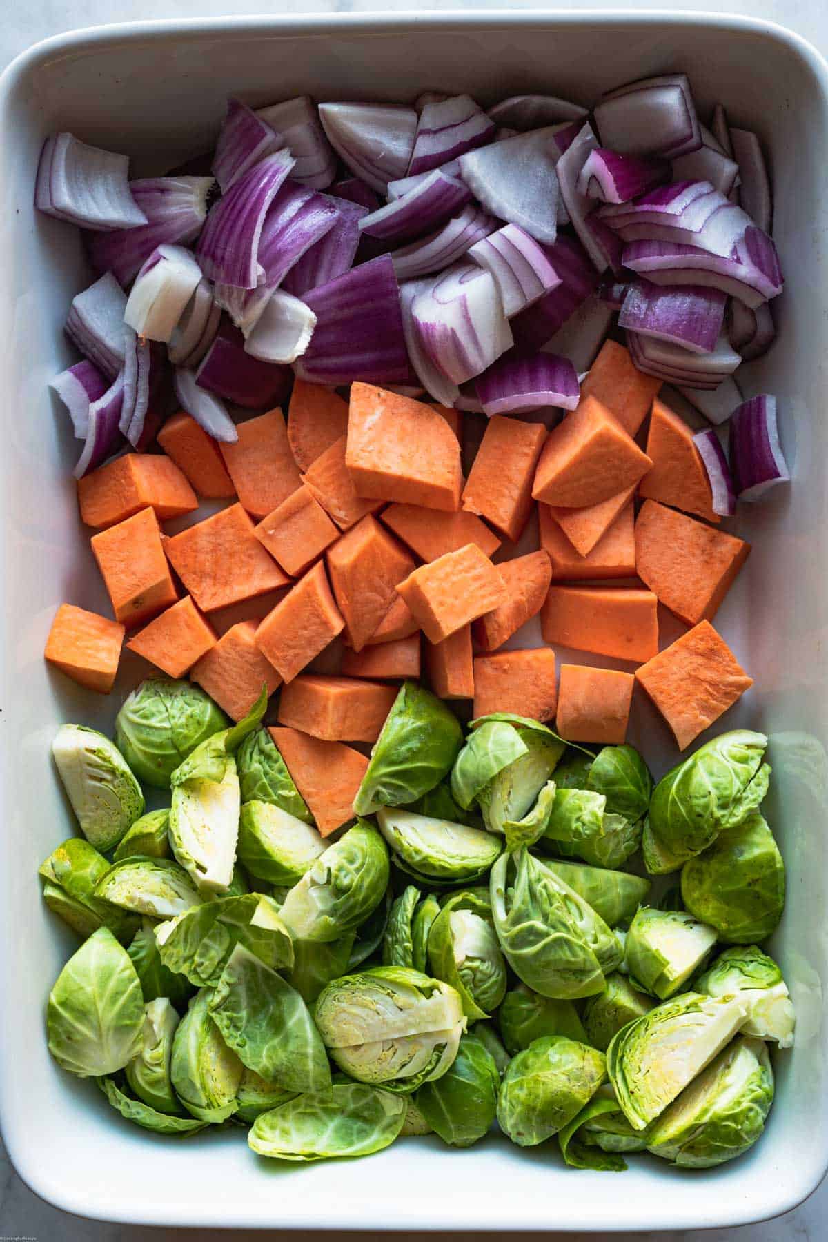 Chopped red onion, sweet potato, and Brussels sprouts in a large casserole dish.