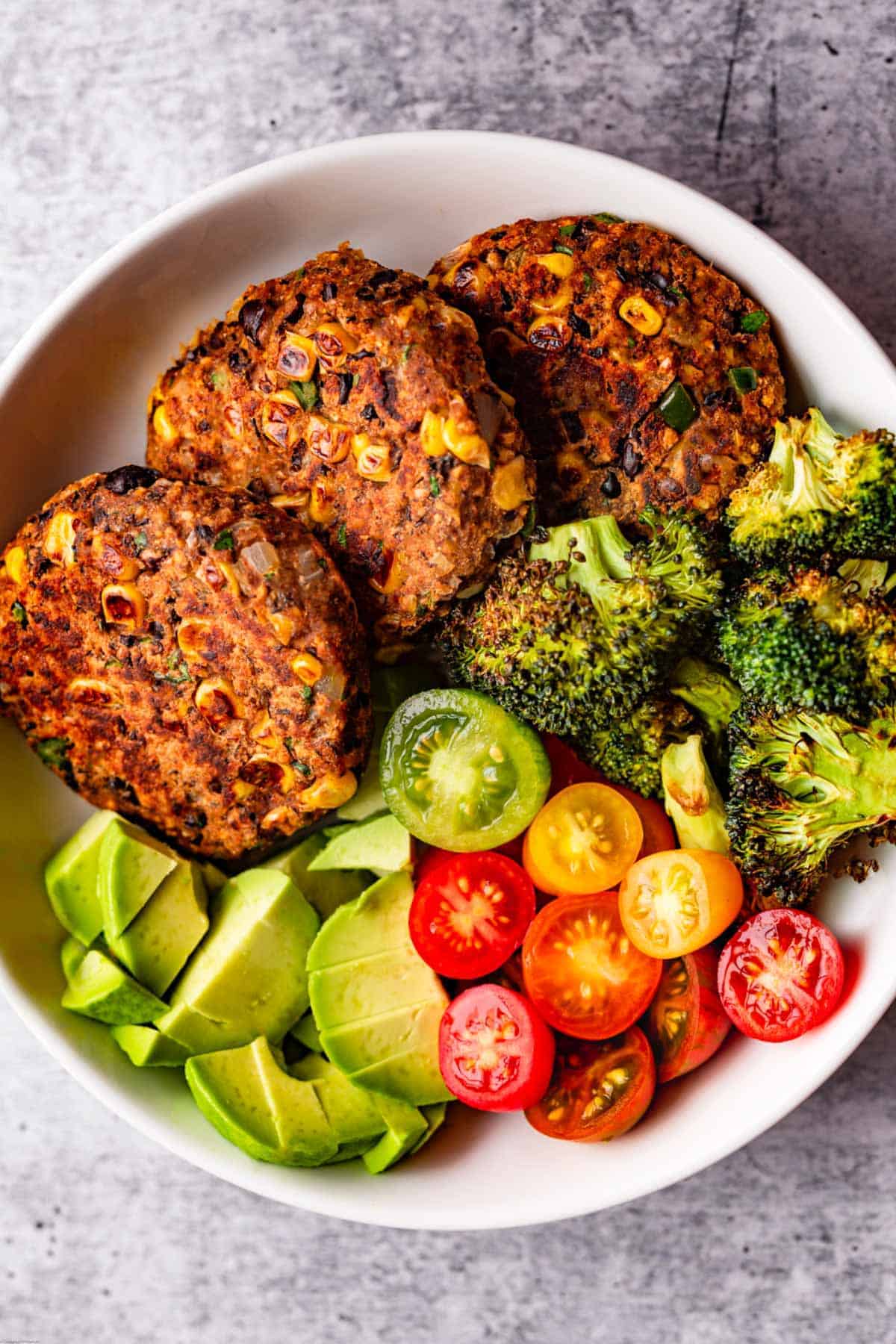 Three meat-like black bean corn burgers, red and yellow sliced cherry tomatoes, chopped avocado, and roasted broccoli in a white bowl.