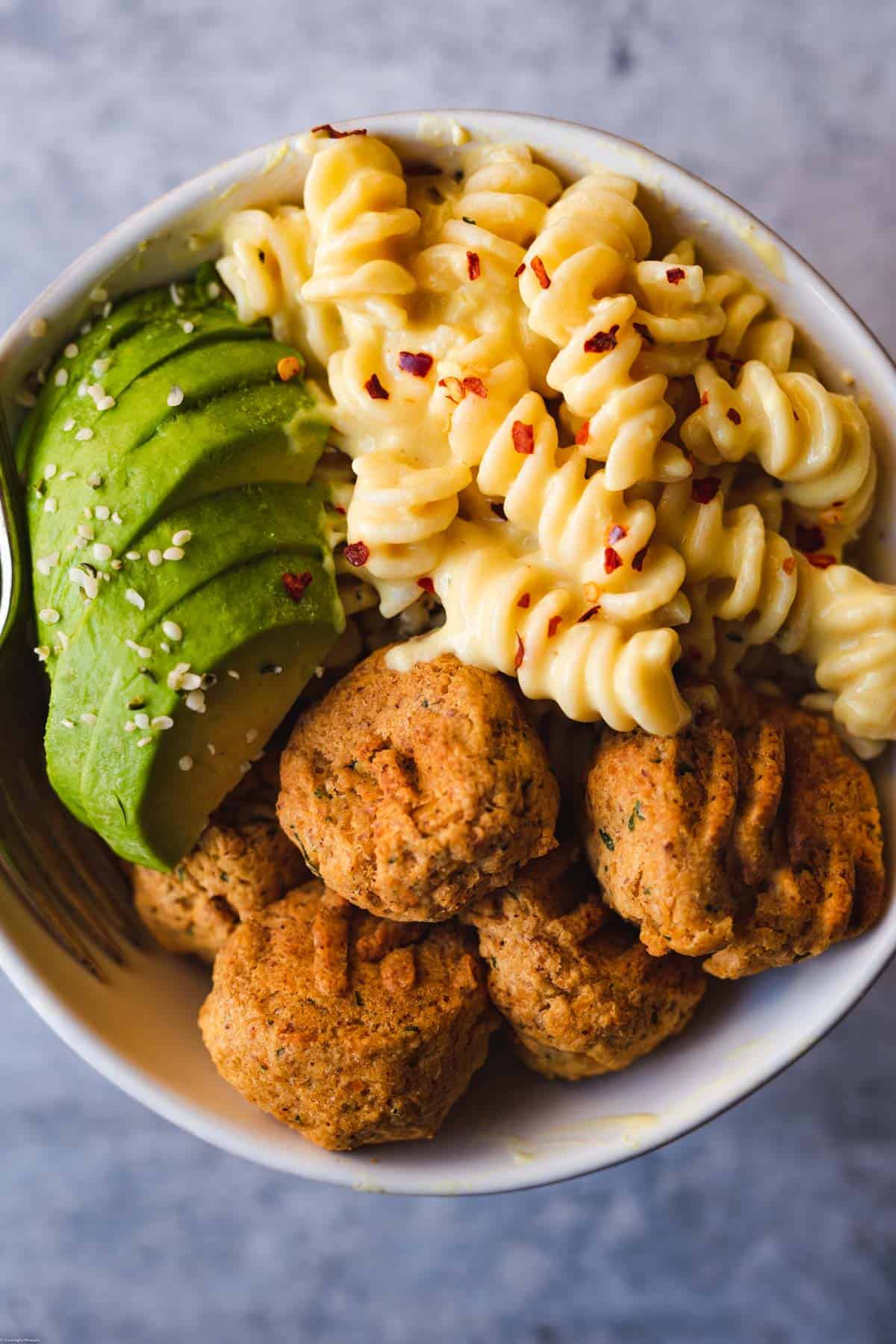 Five golden brown chickpea patties, vegan cheesy pasta, and sliced avocado topped with crushed red pepper in a bowl.