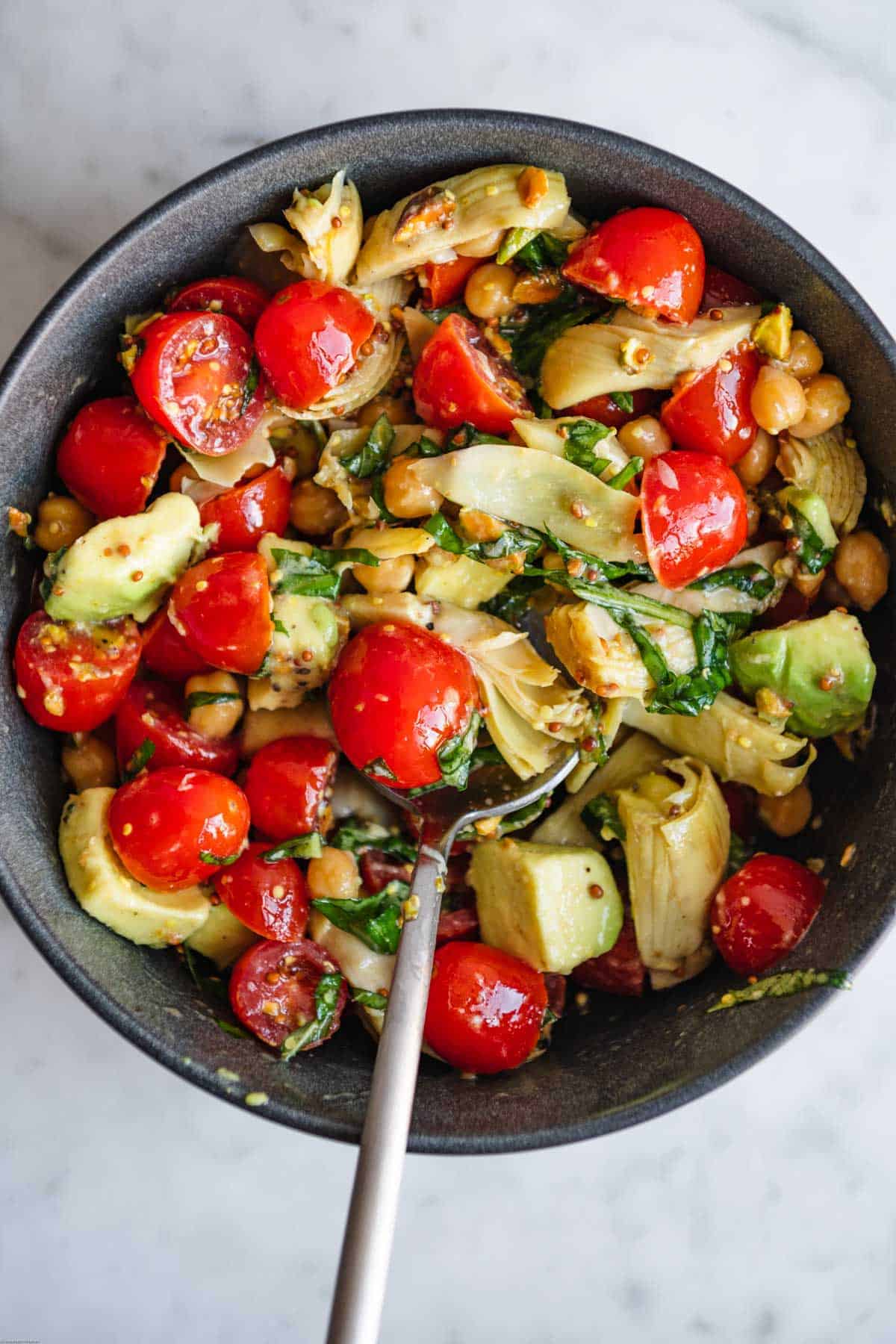 Halved grape tomatoes, avocado chunks, fresh mint and basil, grain mustard dressing, artichoke hearts, chopped pistachios, and chickpeas mixed in a bowl.