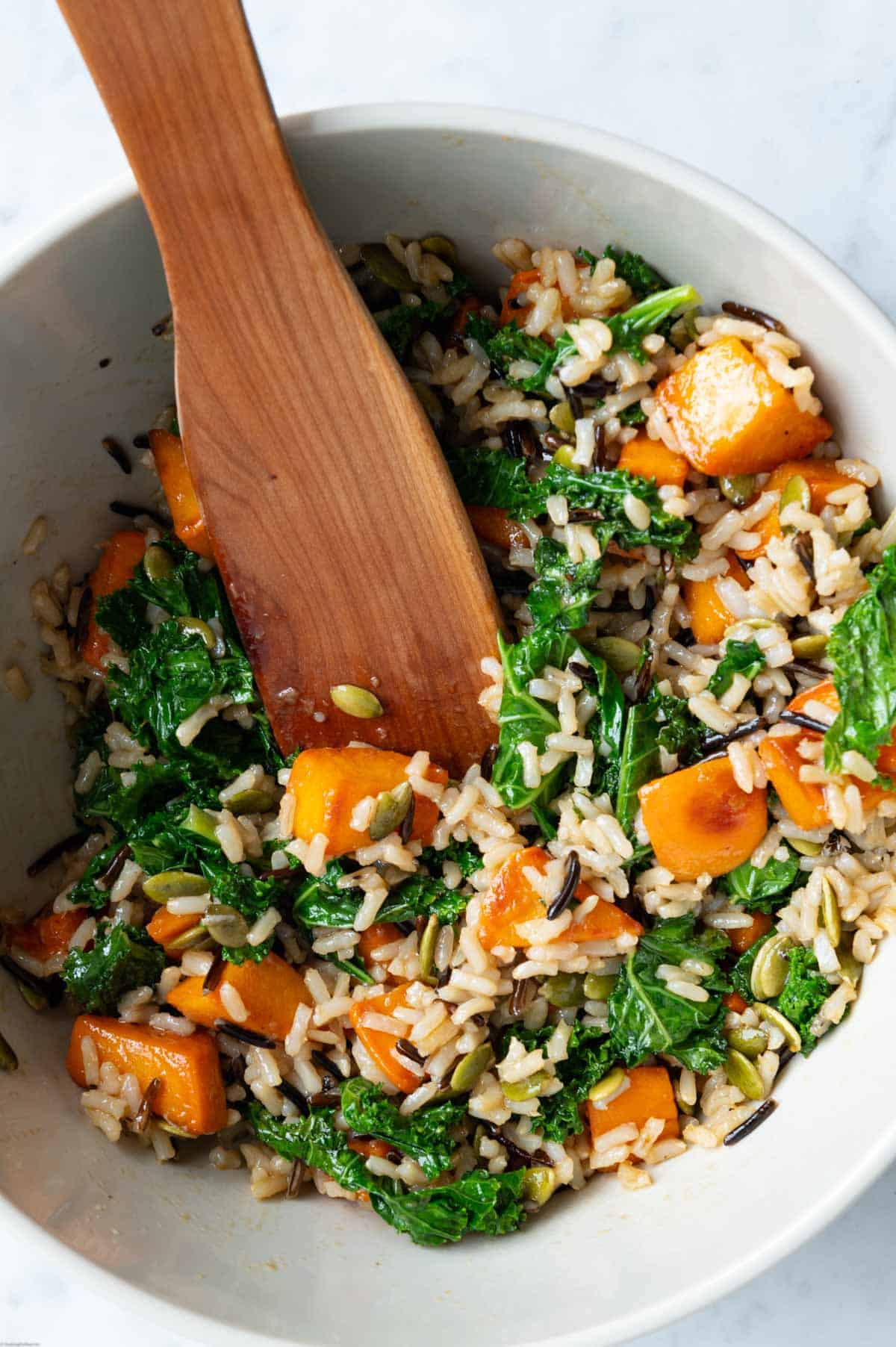 Sweet potato, kale, roasted pepitas, wild rice blend, and hemp hearts mixed with dressing in a white bowl with a wooden spatula.