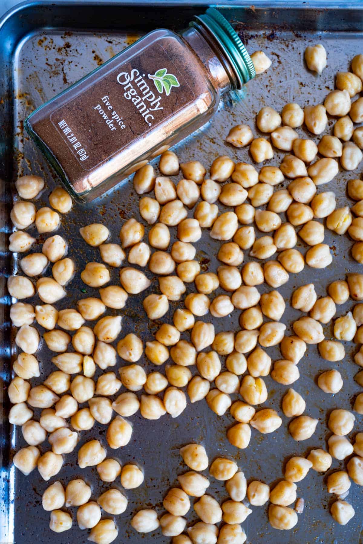 Chickpeas spread out on a stainless steel rimmed baking sheet with olive oil and five spice powder.