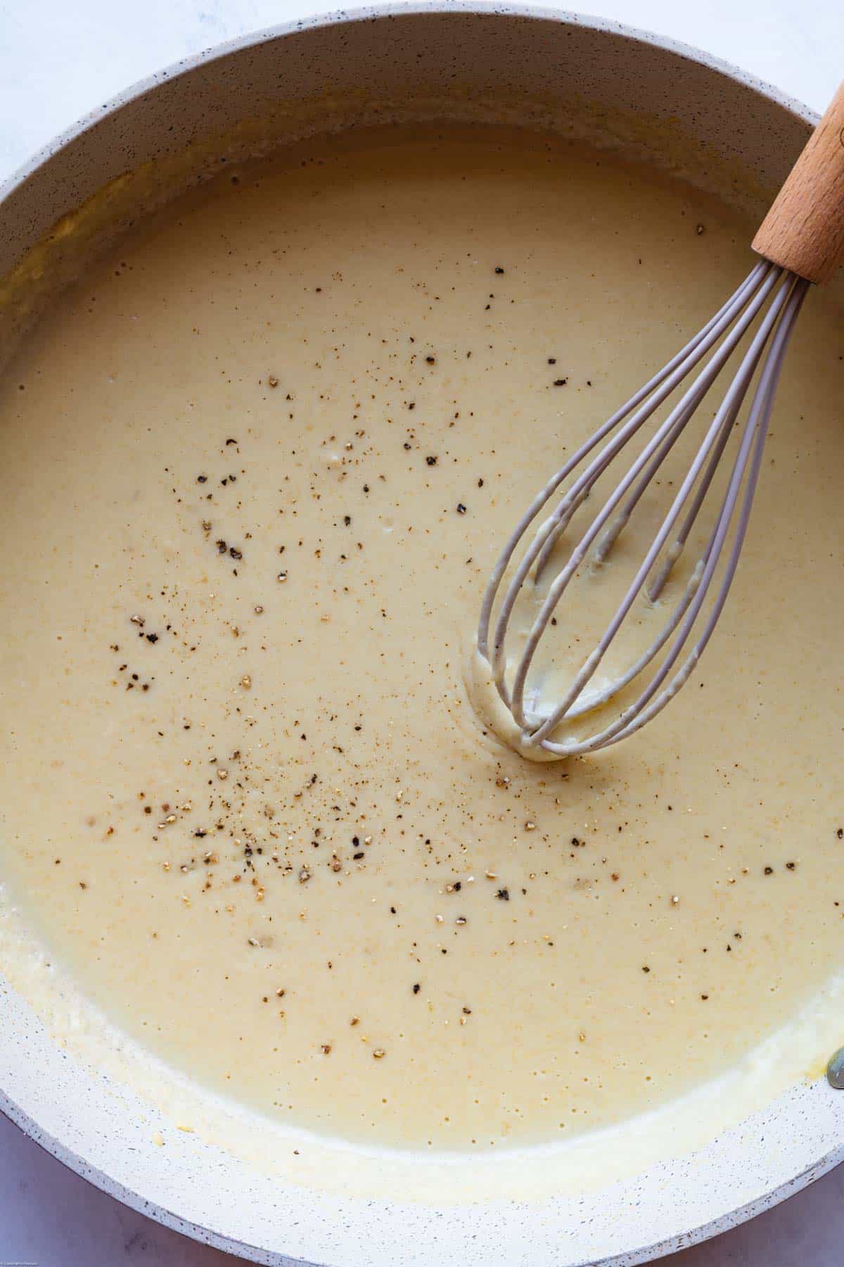 White sauce with black pepper in a large white skillet with a whisk.