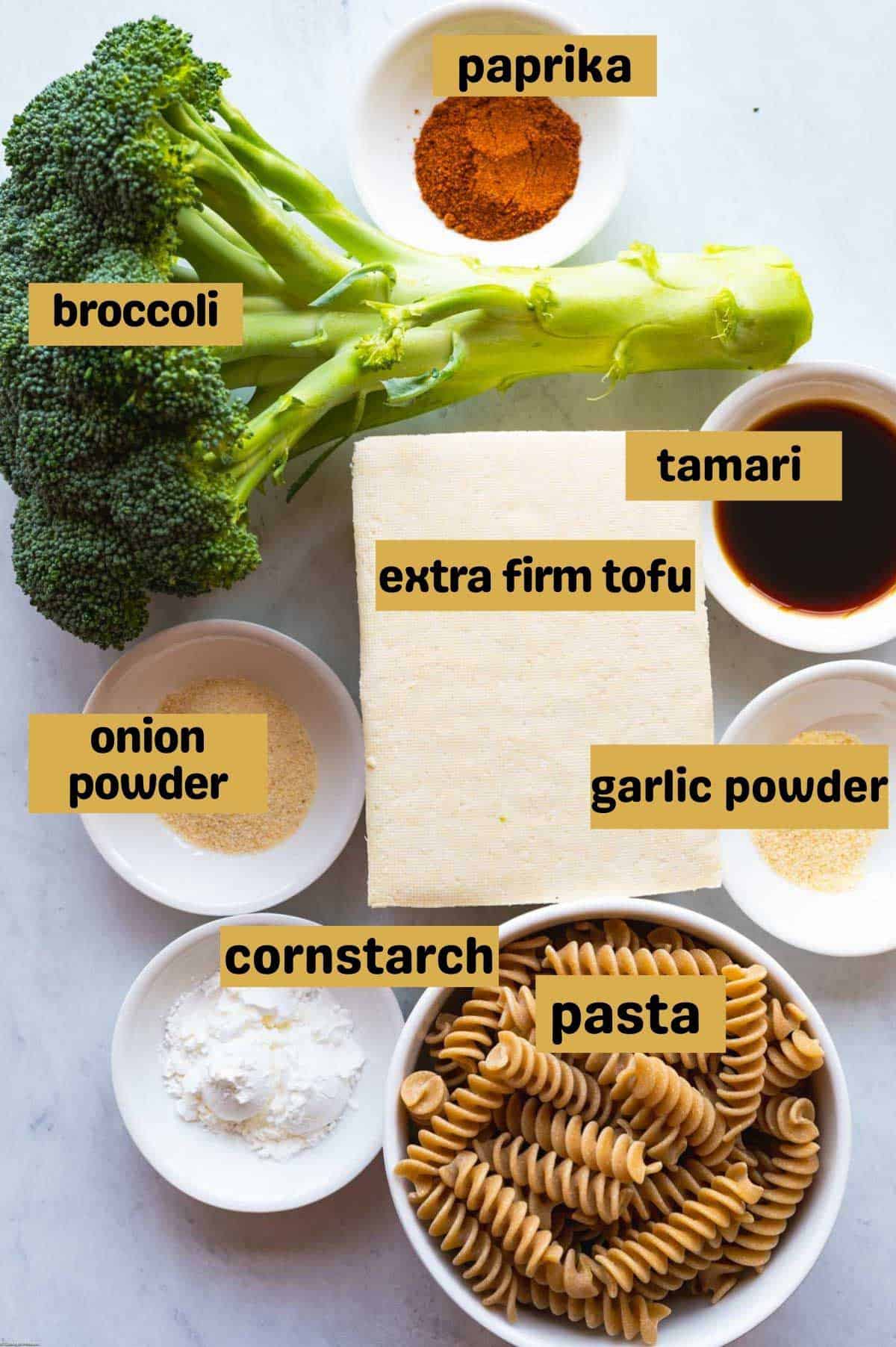 Broccoli, paprika, extra firm tofu, dried pasta, cornstarch, garlic powder, onion powder, and tamari in bowls on a white backdrop.