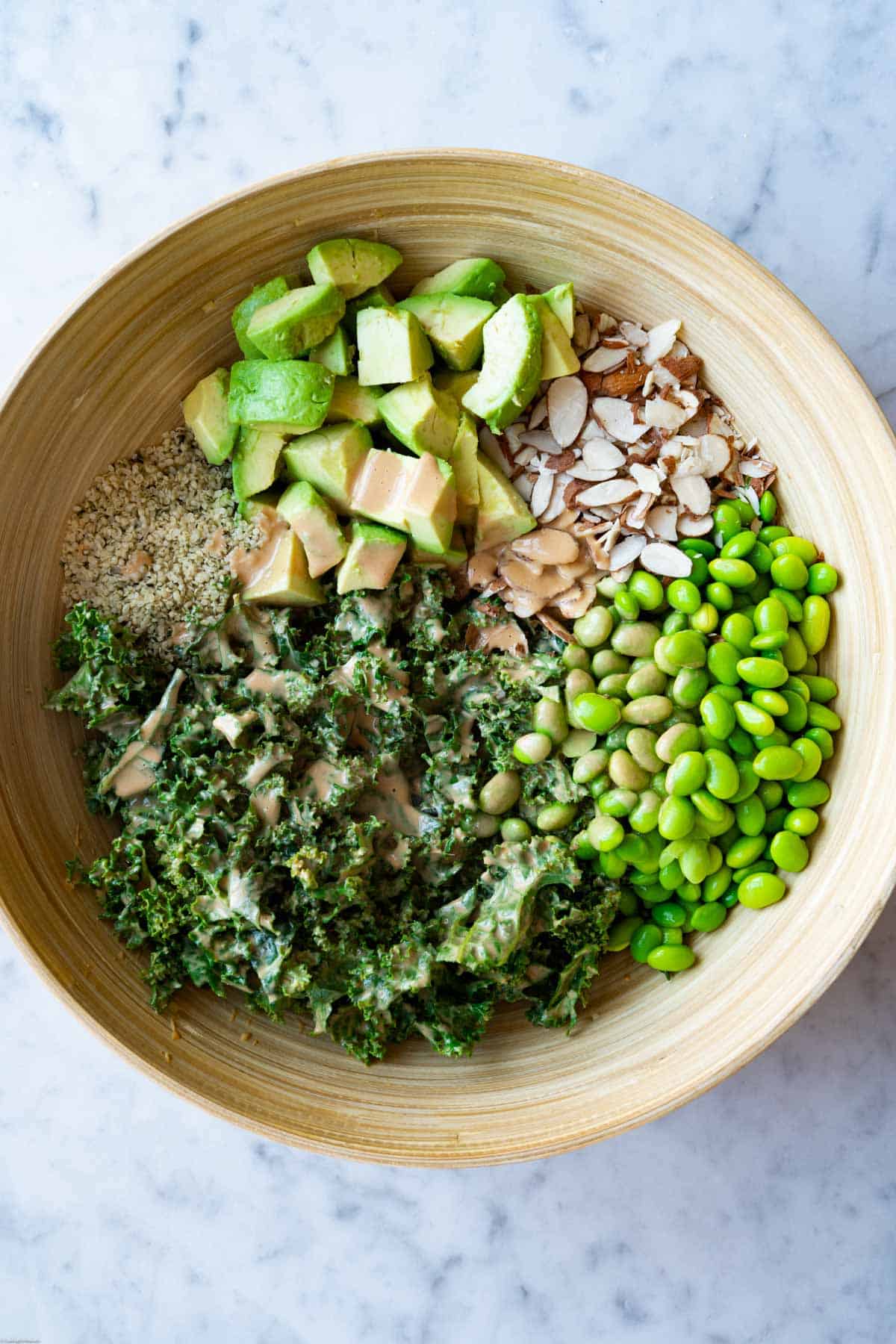 Chopped kale, avocado chunks, slivered almonds, and hemps seeds in a large bamboo bowl, topped with creamy peanut dressing.