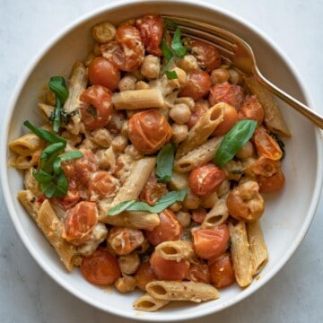 Easy roasted chickpeas, burst tomatoes, creamy hummus, wholewheat pasta, and roasted garlic cloves in a white bowl with a fork. Topped with chopped basil leaves.