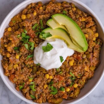 Cooked quinoa with lentils, corn, diced tomatoes, celery, red onions, and carrots in a white bowl. Topped with vegan sour cream, avocado, and chopped cilantro.