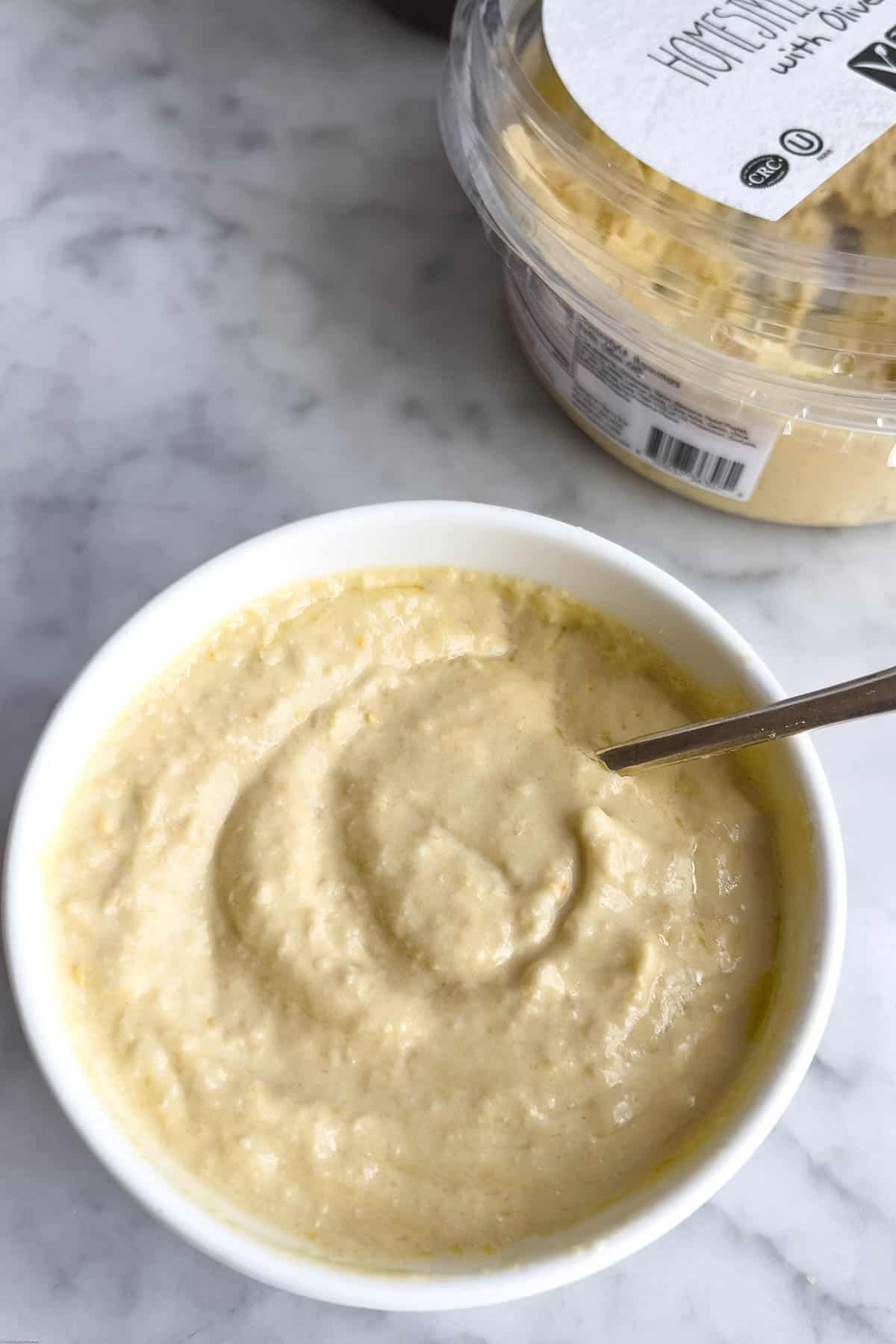 Store-bought hummus in a white bowl with a silver spoon, and a opened container of hummus in the background.