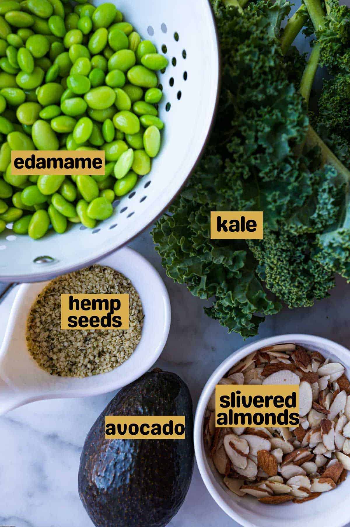Edamame in a white colander, a bunch of kale, hemp seeds in a small white bowl, one avocado, and slivered almonds in a small white bowl. Labeled text. On a white marble tile.