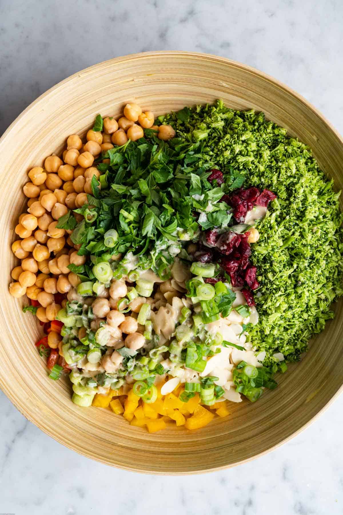 Riced broccoli, chickpeas, diced red and yellow bell pepper, sliced green onion, chopped parsley and mint, sliced almonds, and oil-free tahini dressing in a large wooden bowl.