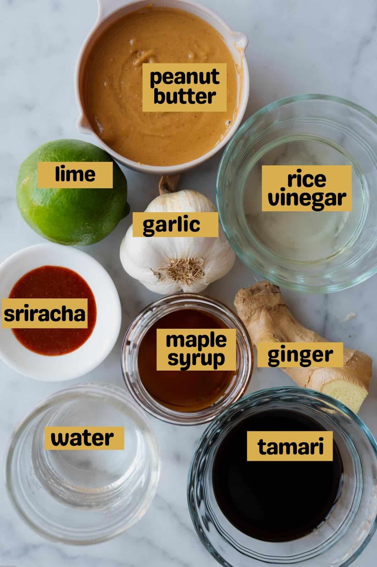 Peanut butter in a white bowl, one lime, one head garlic, one piece of ginger, rice vinegar in a glass bowl, sriracha is a white bowl, maple syrup in a glass bowl, tamari in a glass bowl, and water in a glass bowl. On a white marble tile.