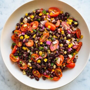 Black beans, quartered grape tomatoes, chopped cilantro, corn kernels, and chopped red onion in a white bowl on a white marble board.
