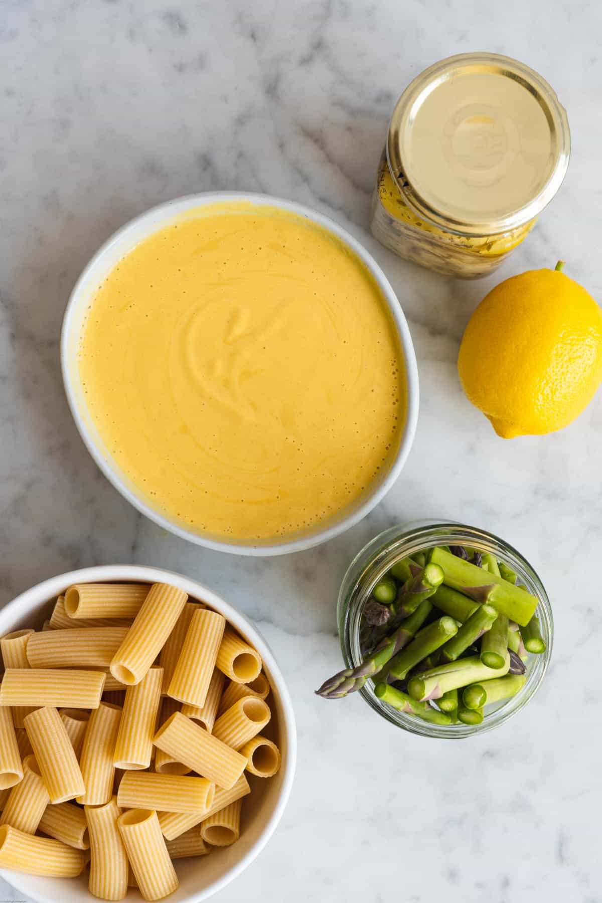 Rigatoni in a white bowl, cut asparagus spears in a glass jar, one lemon, jarred artichoke hearts, and Creamy Dairy-Free Nut-Free Lemon Pasta Sauce in. white bowl.