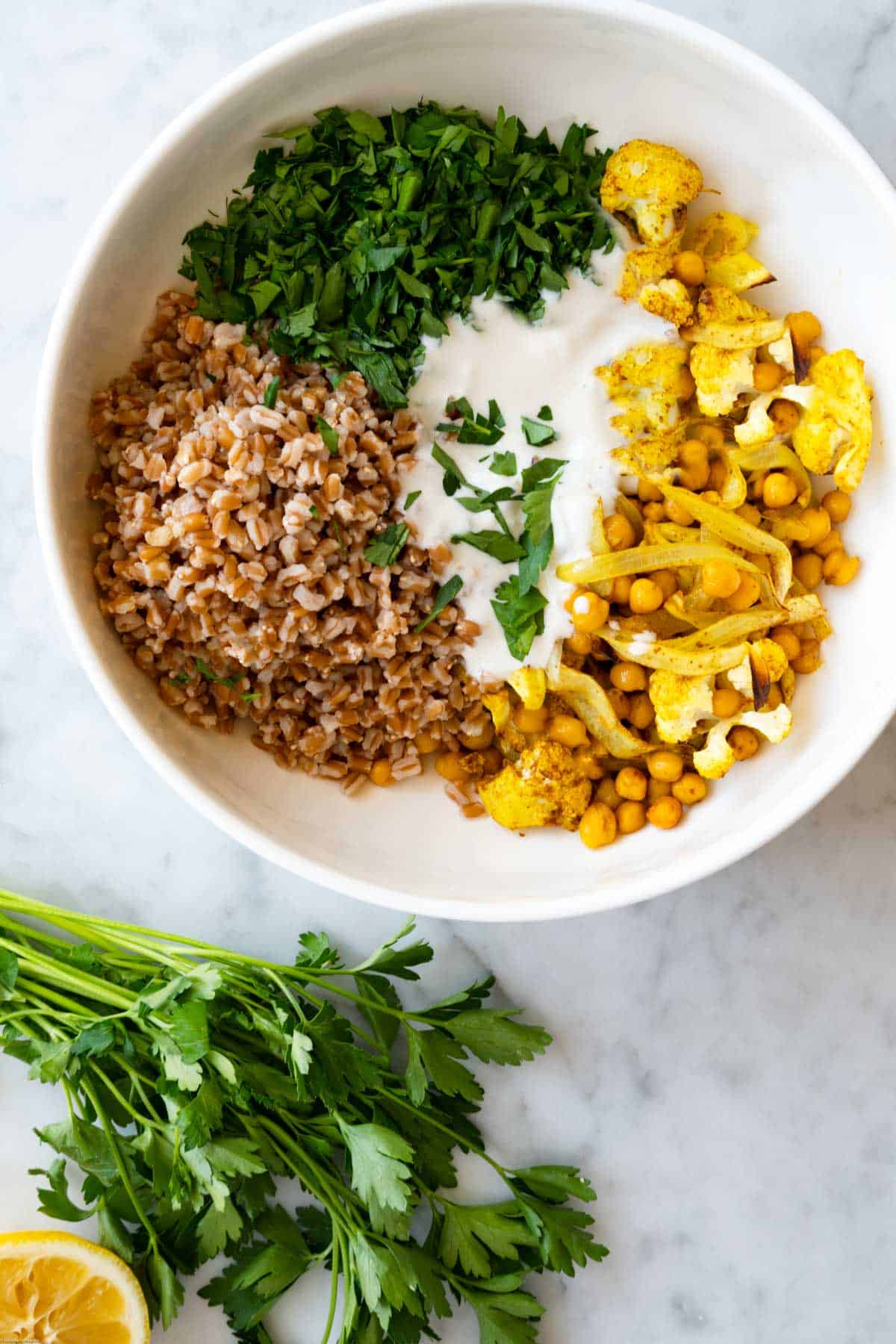 Farro, curry roasted cauliflower, chickpeas, and onion, white yogurt dressing, and chopped parsley in a white bowl on a white marble table.