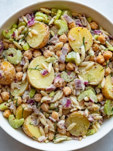 Baby potato and pasta salad with chopped celery, red onion, dill, chickpeas, and creamy tahini and mustard dressing in a white bowl.