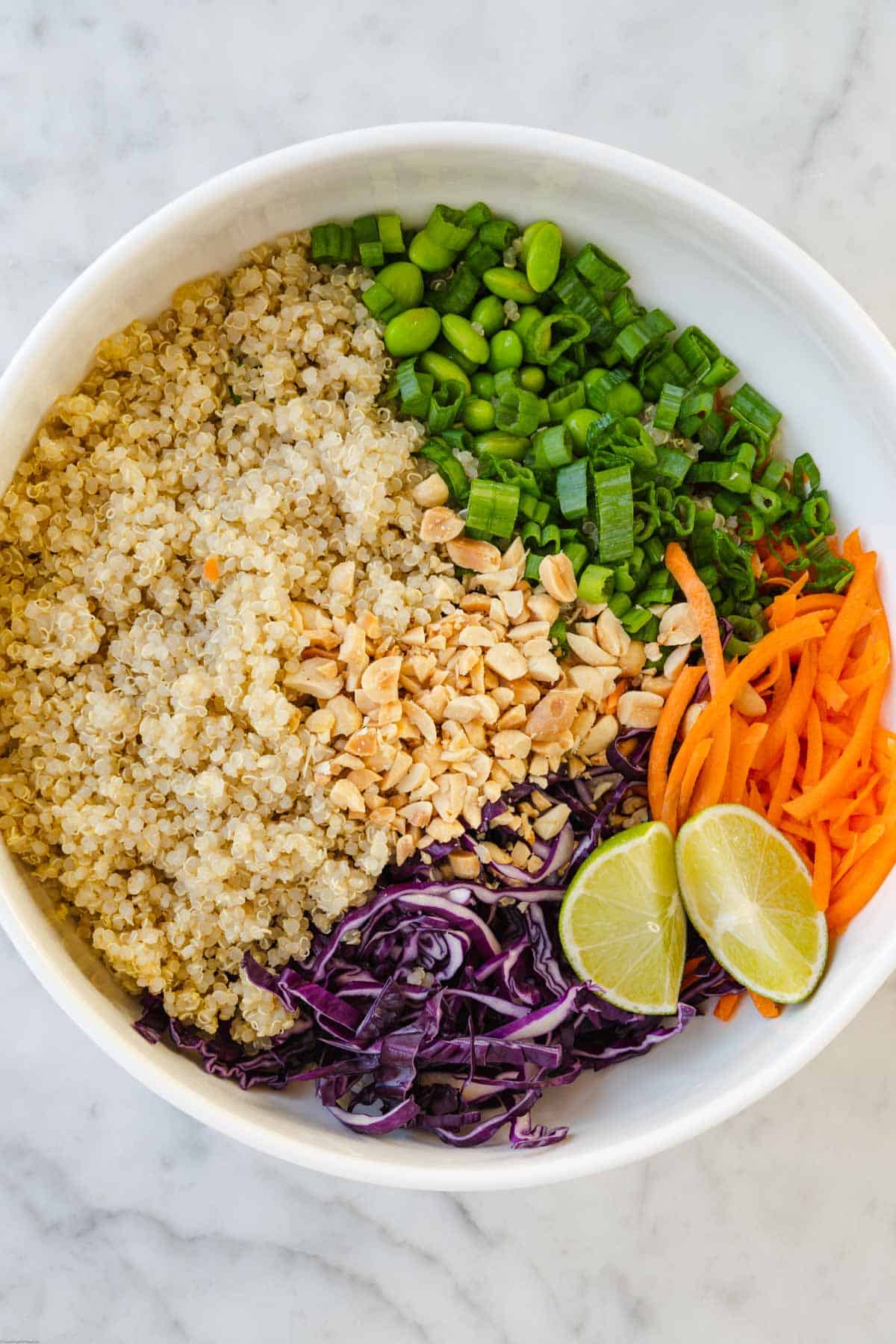 Quinoa, edamame, shredded carrots and red cabbage, chopped green onion, and chopped peanuts in a white bowl with two lime wedges.