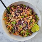 Quinoa, edamame, shredded carrots and red cabbage, chopped green onion, and chopped peanuts mixed together in a white bowl with a black fork. Served with lime wedges.