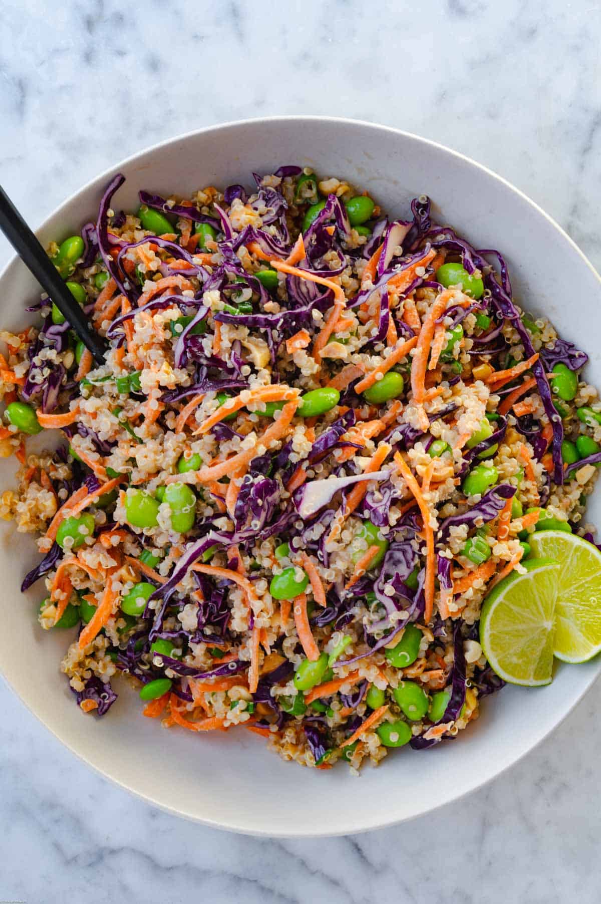 Quinoa, edamame, shredded carrots and red cabbage, chopped green onion, and chopped peanuts mixed together in a white bowl with a black fork. Served with lime wedges.