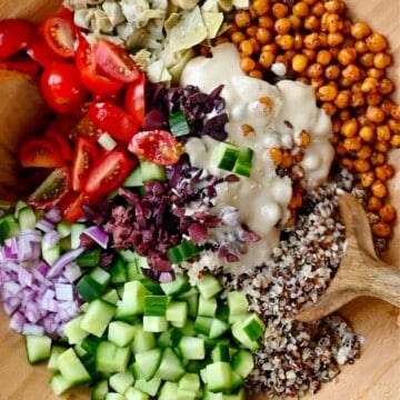 Quinoa, arugula, roasted chickpeas, chopped cucumber, red onion, grape tomatoes, kalamata olives, artichoke hearts, and creamy hummus dressing in a wooden bowl.