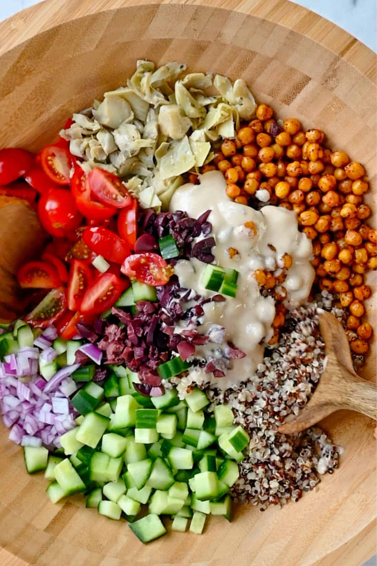 Quinoa, arugula, roasted chickpeas, chopped cucumber, red onion, grape tomatoes, kalamata olives, artichoke hearts, and creamy hummus dressing in a wooden bowl.