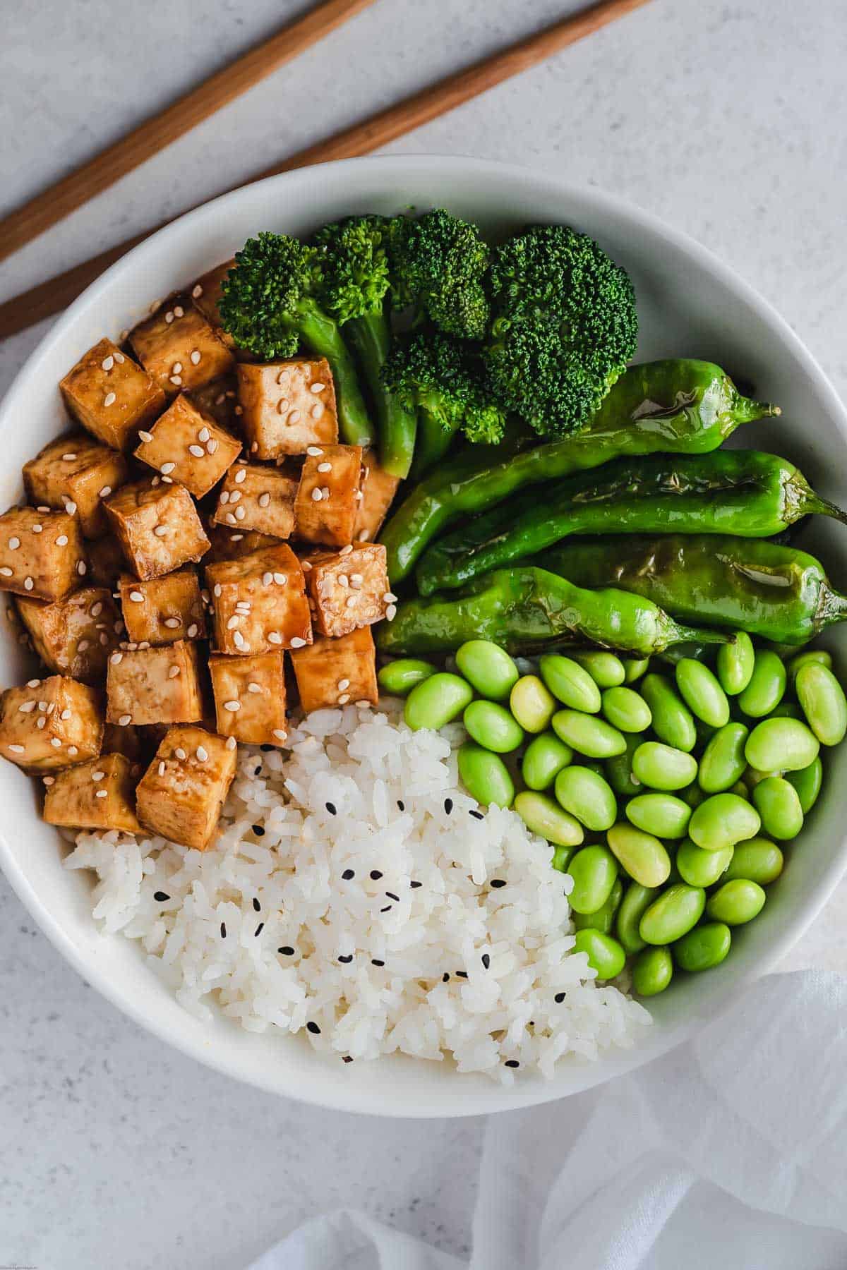 Crispy tofu cubes garnished with sesame seeds, white rice, shelled edamame, broccoli florets, and shishito peppers in a white bowl with bamboo chopsticks.