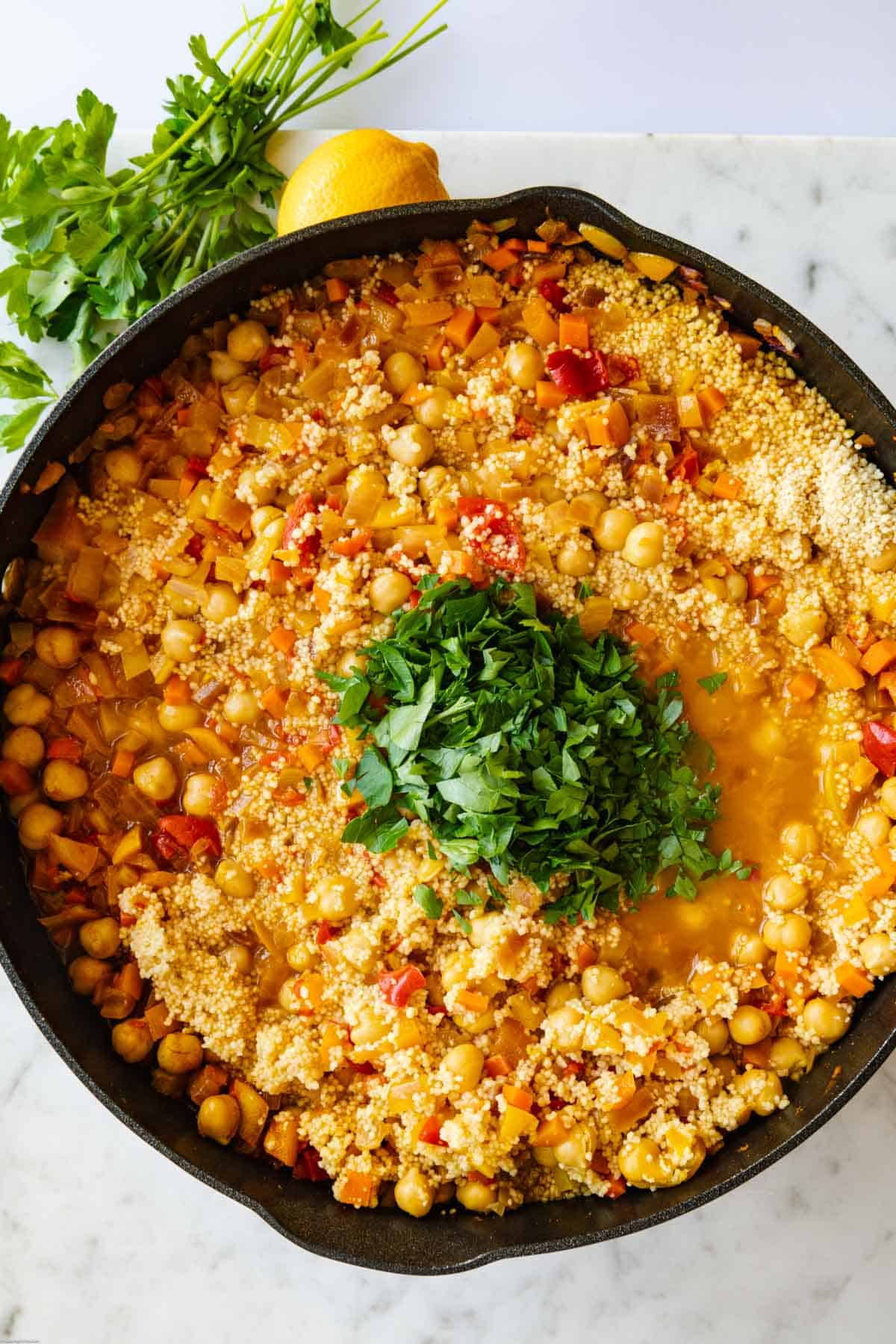 Spiced Moroccan couscous and chickpeas with chopped bell peppers, carrots, red onion, Peppadew piquante peppers, veggie broth, and parsley in a cast iron skillet.
