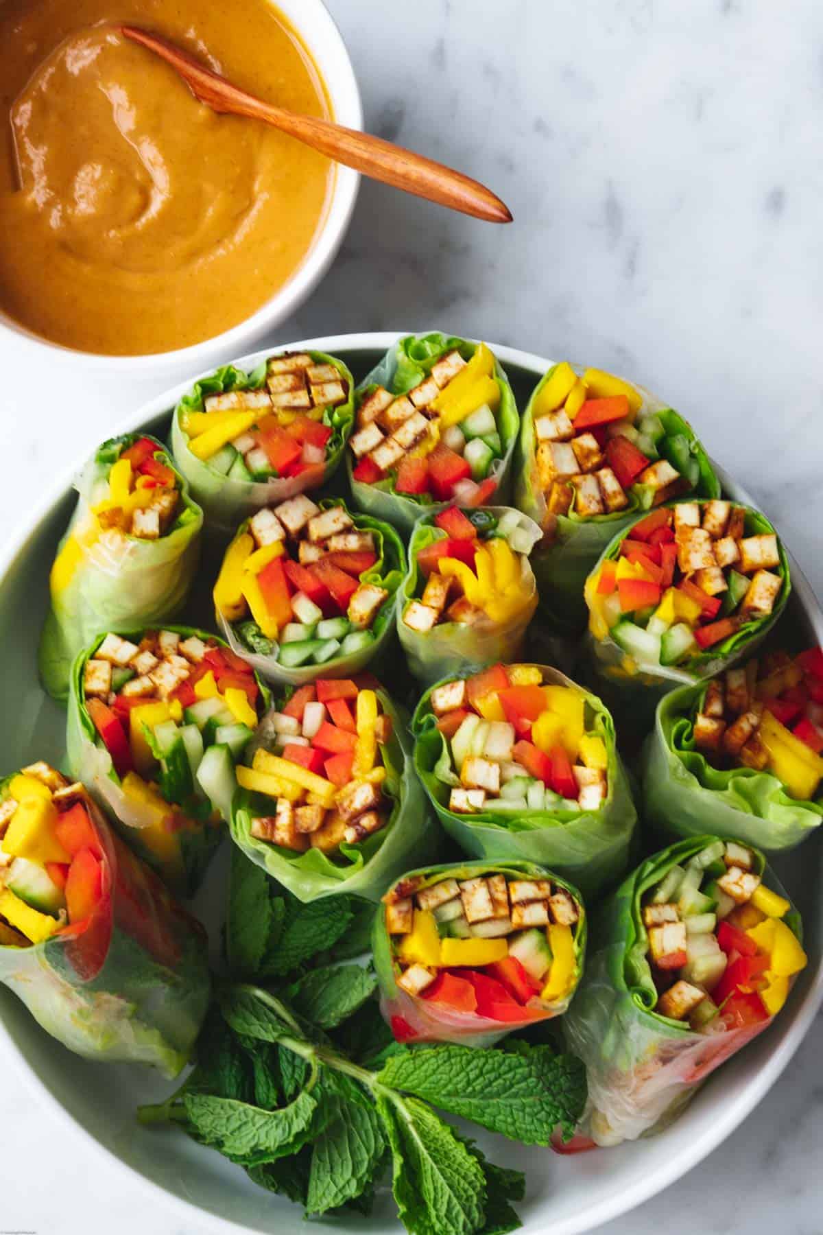 7 summer rolls cut in half in a white bowl. Filled with tofu, mango, cucumber, red bell pepper, lettuce, and mint. Served with peanut dipping sauce.