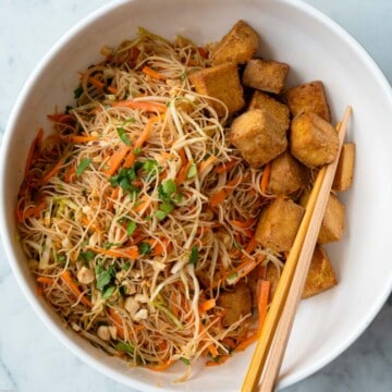 A salad with shredded carrots and green cabbage, chopped peanuts, chopped cilantro, vermicelli noodles, and air fryer tofu cubes, in a white bowl with bamboo chopsticks.