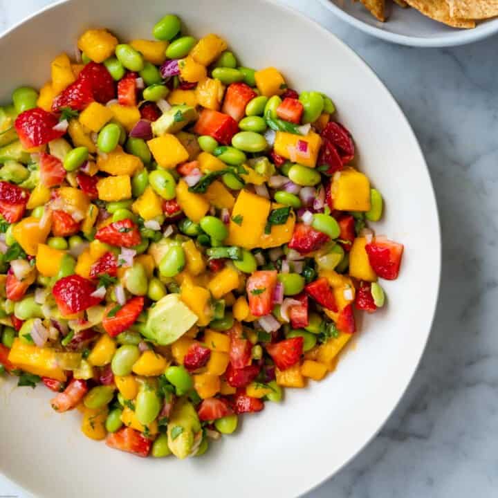 Salsa with mango, strawberries, avocado, edamame, red onion, basil, cilantro, and lime dressing in a white bowl with a side of tortilla chips.