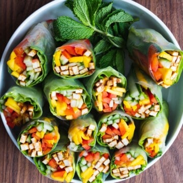 7 summer rolls cut in half in a white bowl. Filled with tofu, mango, cucumber, red bell pepper, lettuce, and mint. Served with peanut dipping sauce.