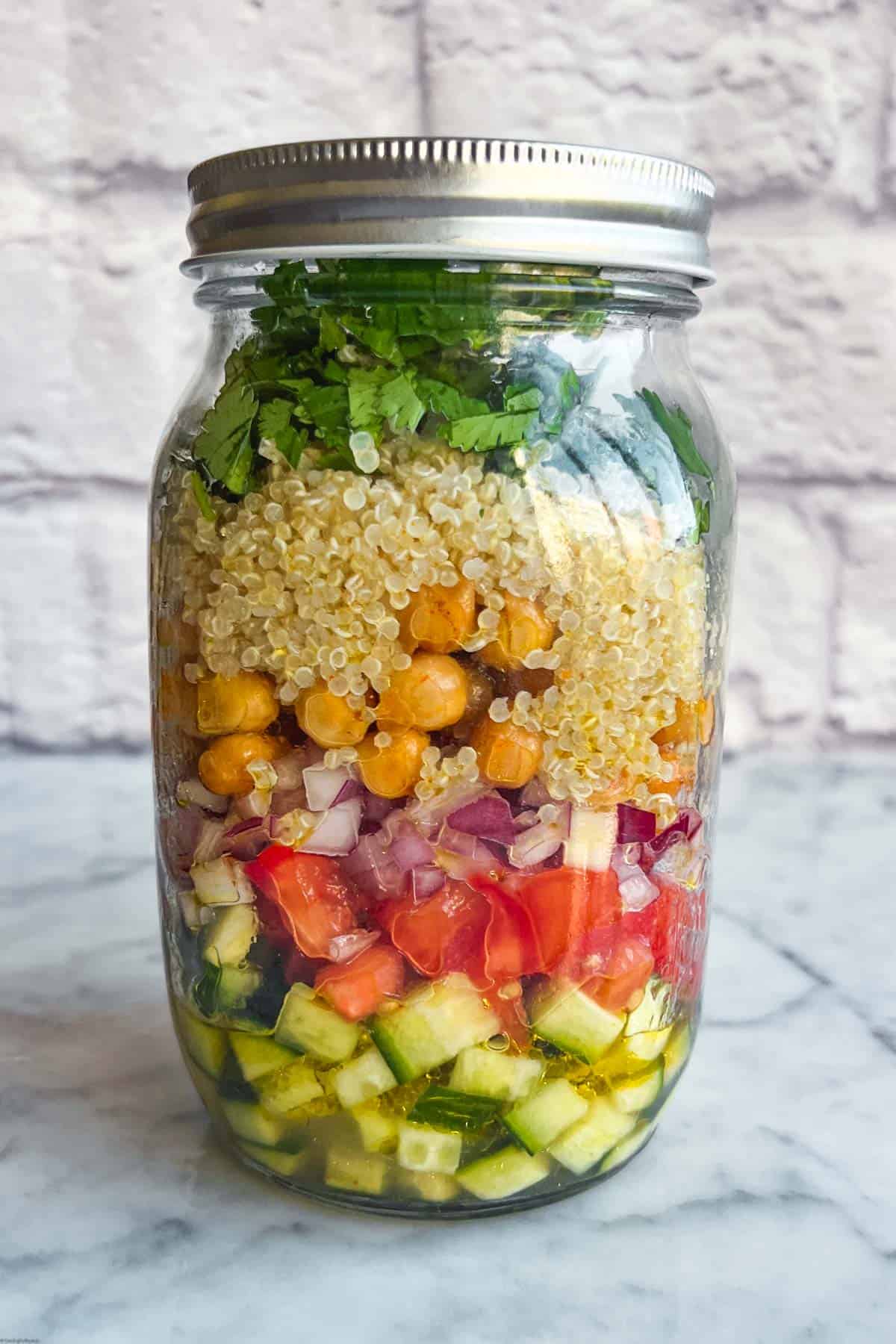 32-ounce salad jar with olive oil, lemon juice, chopped cucumber, tomato, red onion, roasted chickpeas, quinoa, hemp seeds, and cilantro.