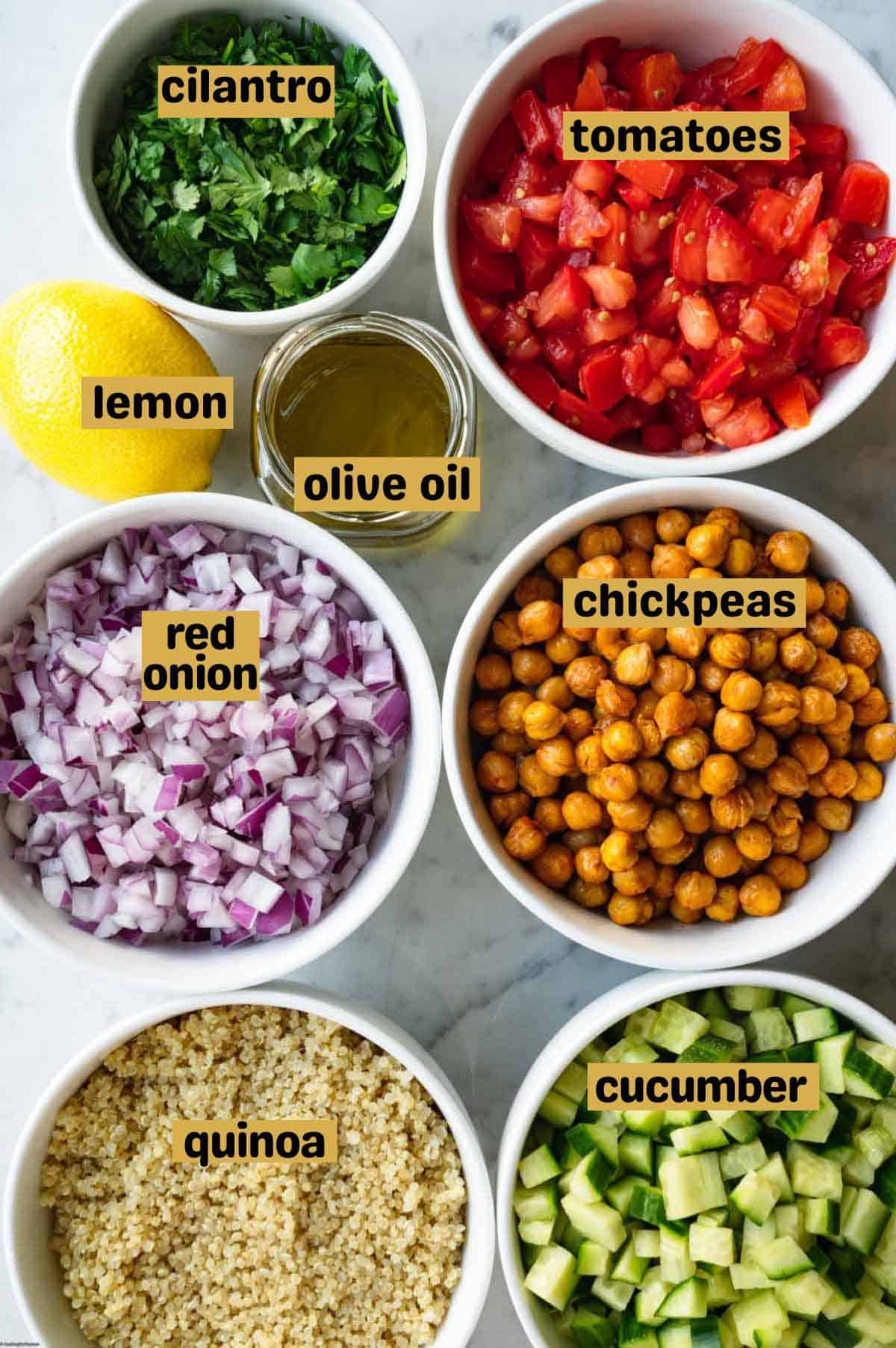 Olive oil in jar, chopped cucumber, tomato, red onion, cilantro, roasted chickpeas, and quinoa, in white bowls, and a lemon.