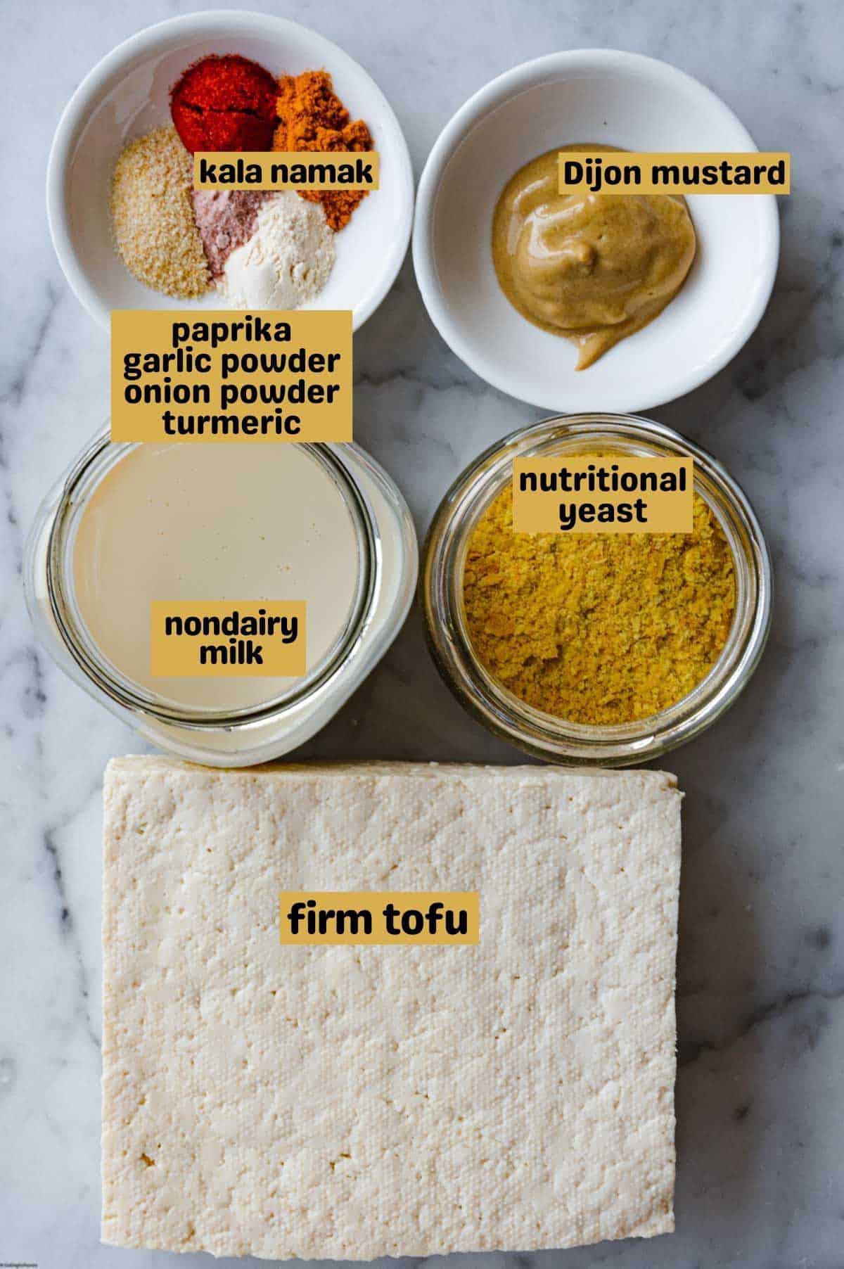 Firm tofu, Dijon mustard in a white bowl, nondairy milk and nutritional yeast in glass jars, and paprika, garlic powder, onion powder, turmeric, and kala namak in a white bowl. On a white marble backdrop.