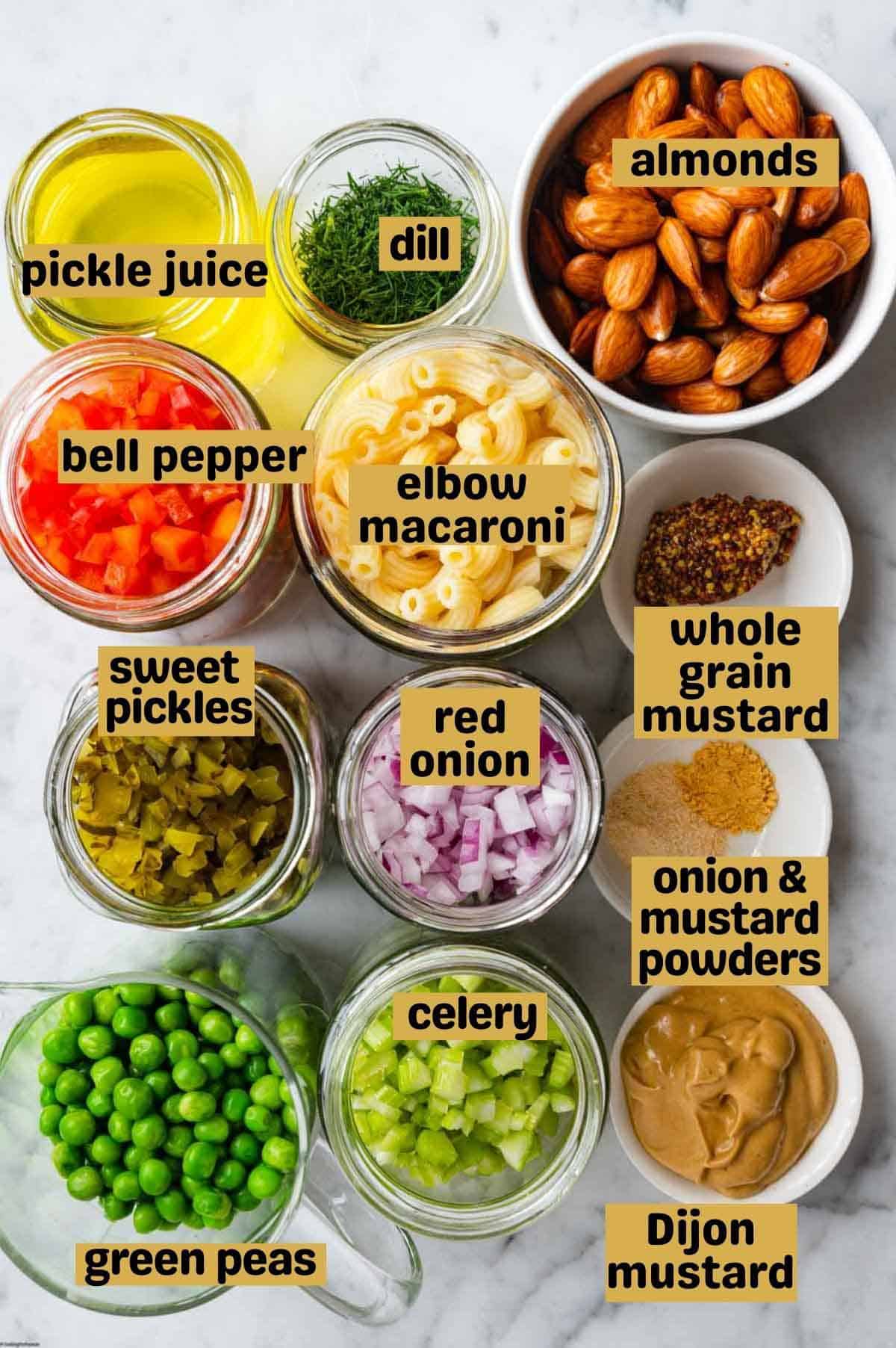 Pickle juice, dill, almonds, chopped red bell pepper, elbow macaroni, whole grain mustard, chopped sweet pickles, chopped red onion, onion and mustard powder, green peas, chopped celery, and Dijon mustard in separate containers on a white marble board.