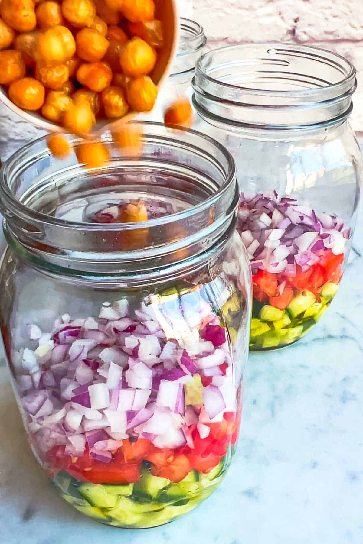 Adding roasted chickpeas to three 32-ounce salad jars with olive oil, lemon juice, chopped cucumber, tomato, and red onion.