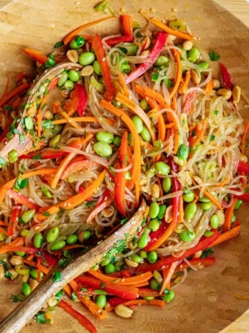 Glass noodle salad with chopped red bell peppers, carrots, cilantro, scallions, peanuts, edamame, sesame seeds, and red chili pepper mixed in a wooden bowl.
