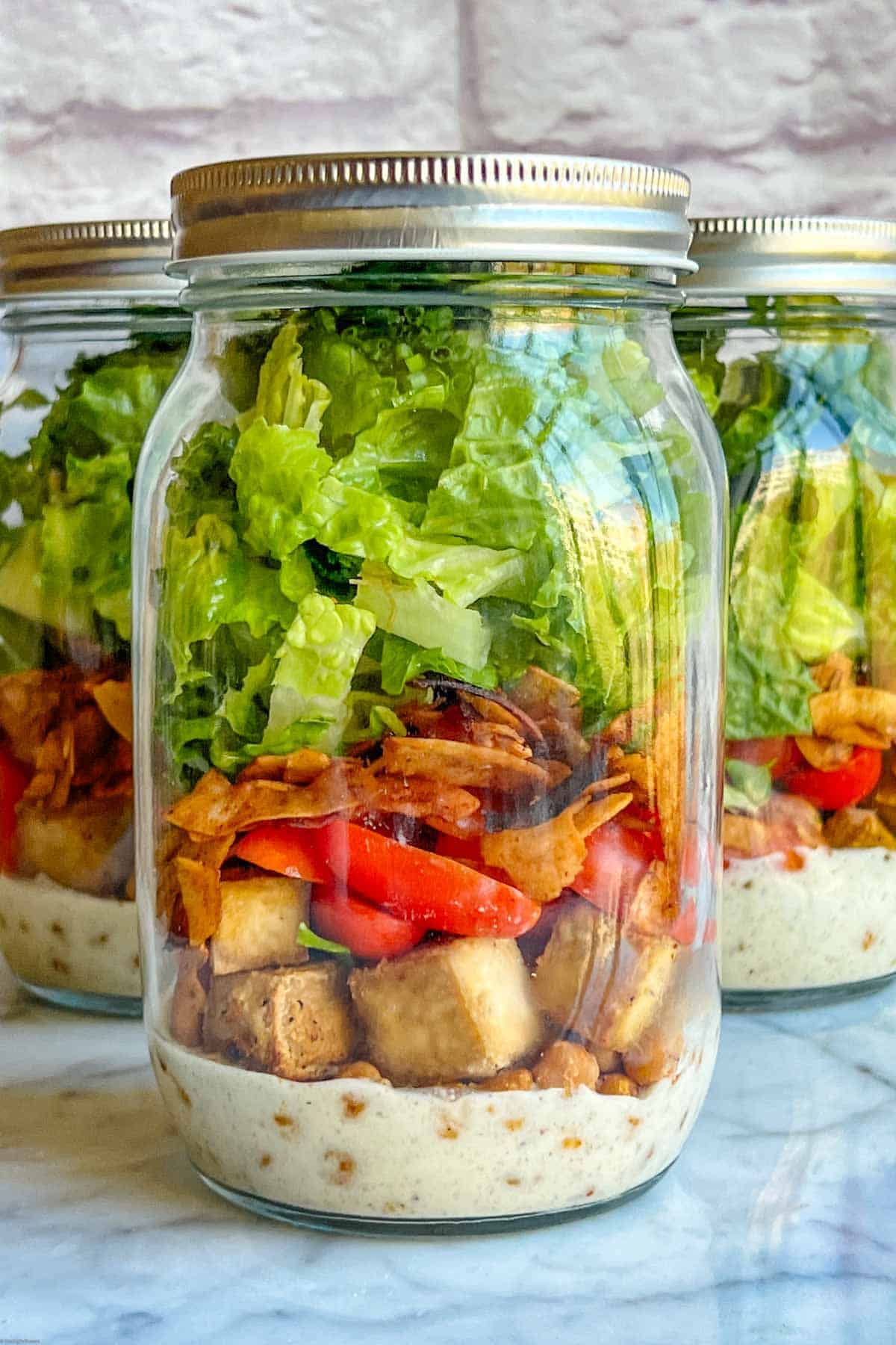 Vegan Cobb Salad with Romaine, halved grape tomatoes, chickpeas, coconut bacon, and white hemp heart dressing in 3 mason jars.