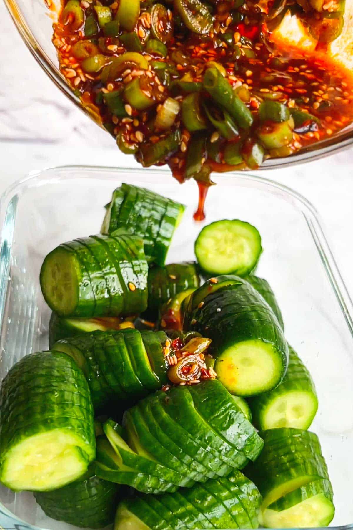 Mini cucumbers cut into accordions in a glass dish with red Korean Gochugaru dressing being added.