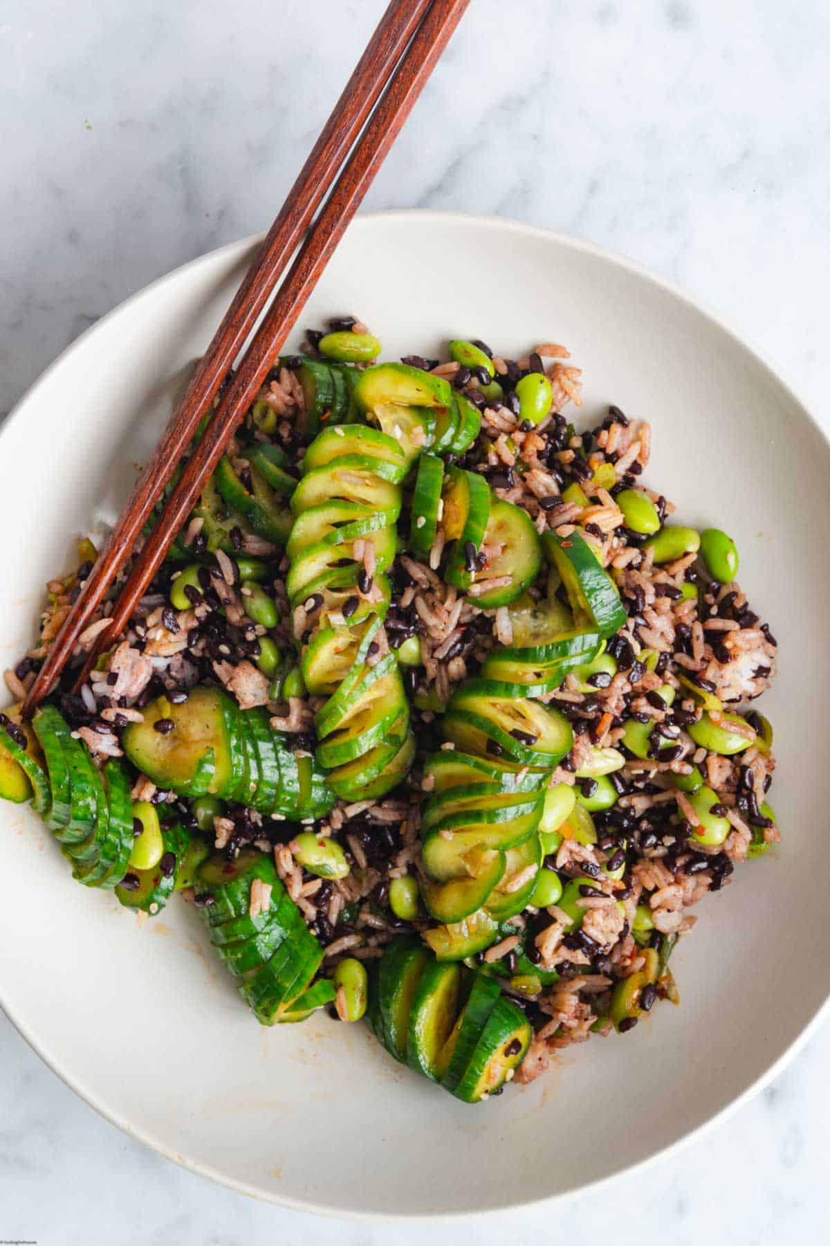 Accordion-cut cucumbers is a red Korean salad dressing with black and white rice, edamame, and chopsticks in a white bowl.