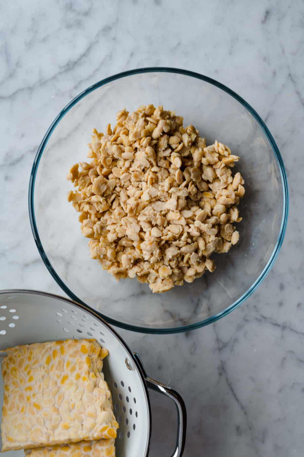 Tempeh crumbles in a glass bowl and a block of tempeh in a white colander.