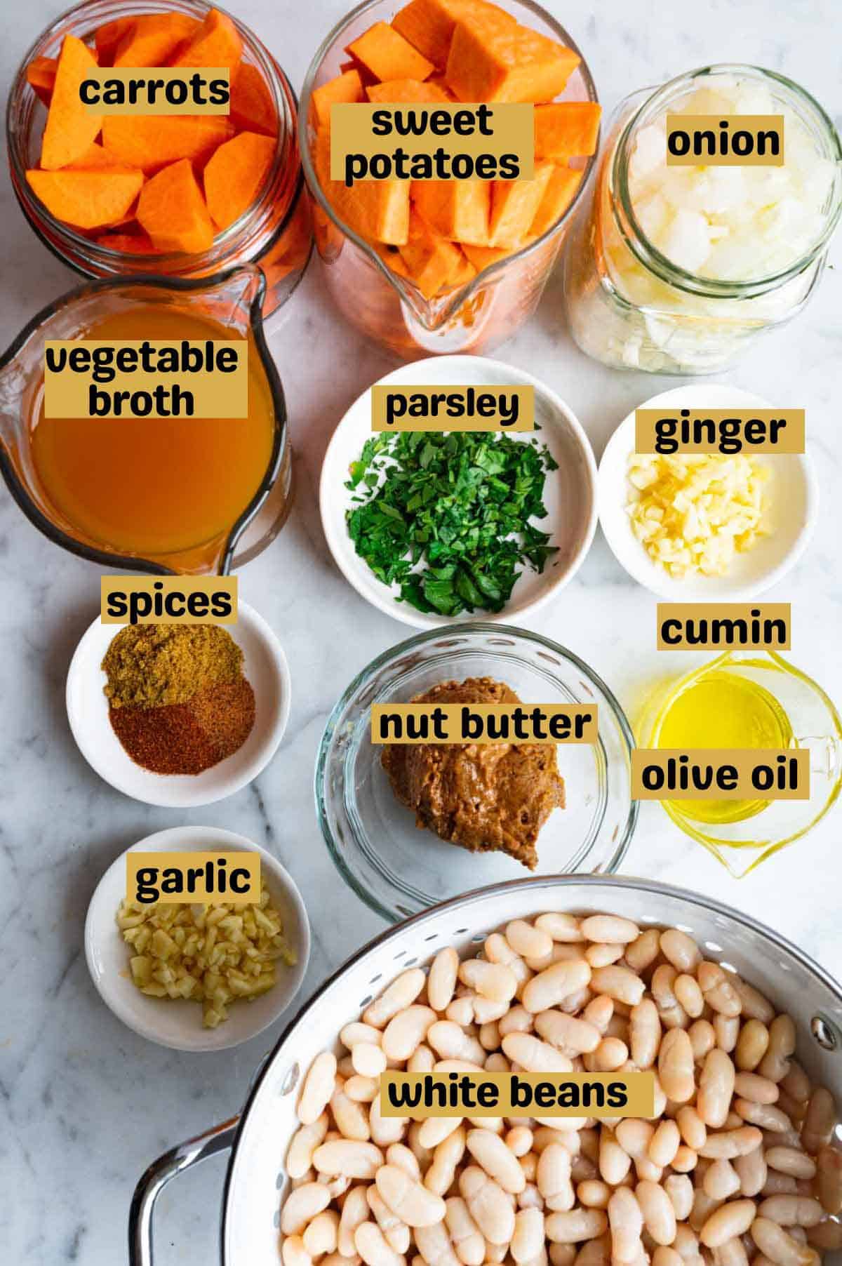 Chopped carrots, sweet potato, onion, garlic, ginger, and parsley, spices, nut butter, olive oil, and vegetable broth in glass jars with a colander of white beans.