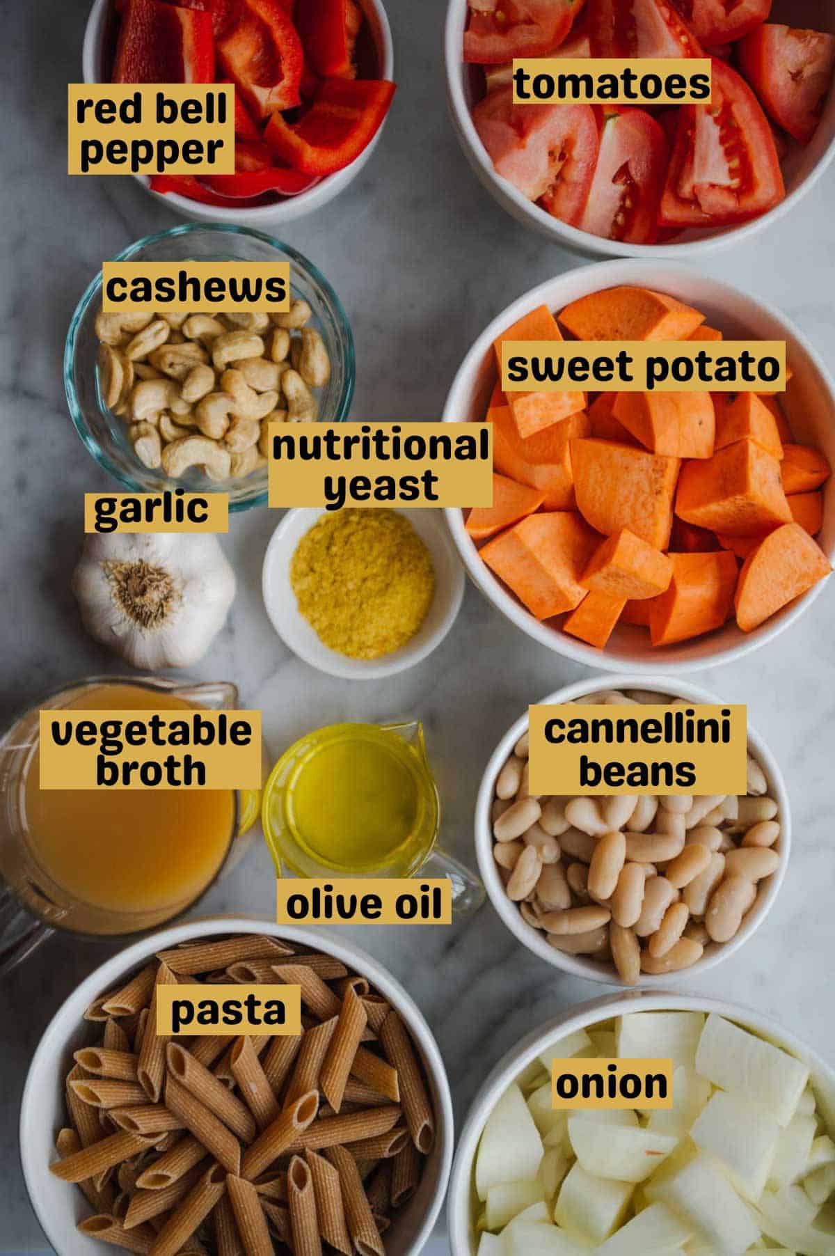 Chopped onion, sweet potato, red bell pepper, tomatoes, penne pasta, cannellini beans, vegetable broth, olive oil, nutritional yeast, garlic bulb, and raw cashews in glass bowls on a white marble board.