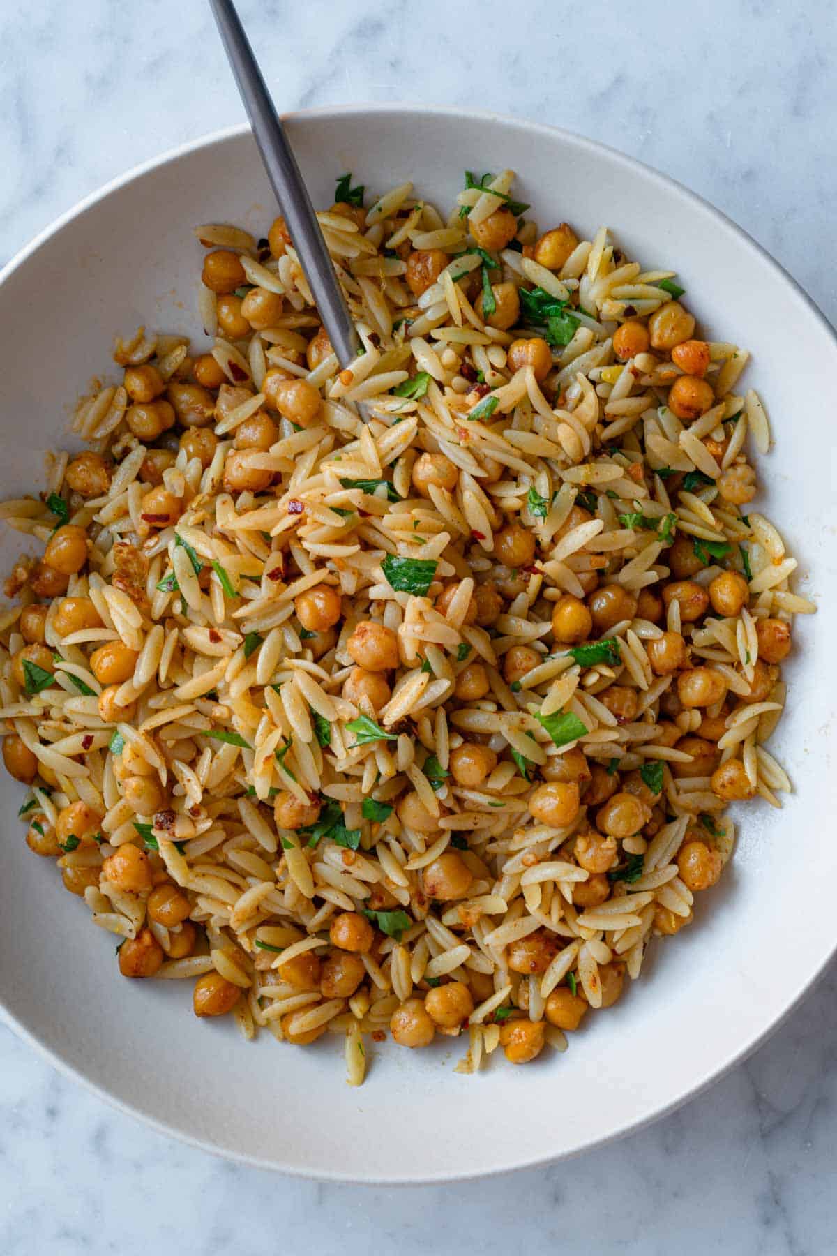 Orzo pasta with roasted chickpeas, chopped parsley, crushed red pepper and lemon zest in a white bowl, with a silver fork.