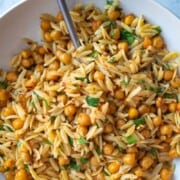 Orzo pasta with roasted chickpeas, chopped parsley, crushed red pepper and lemon zest in a white bowl, with a silver fork.