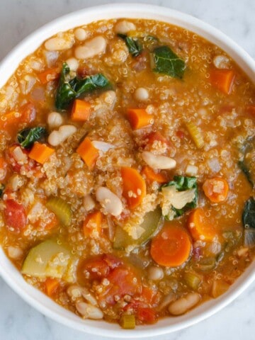 Orange colored vegetable soup with white beans, quinoa, celery, carrots, zucchini, kale, and diced tomatoes in a white bowl.