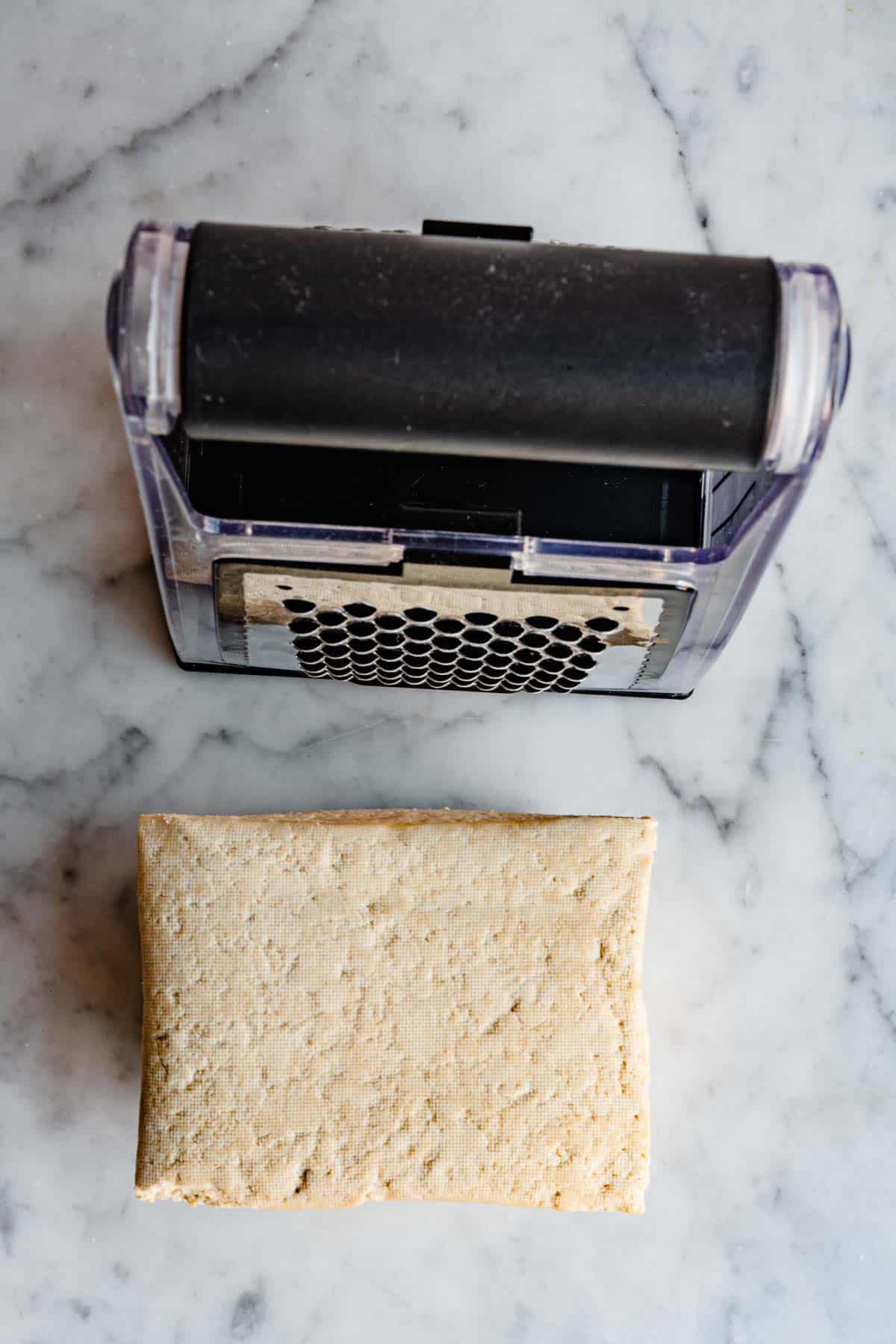 A white block of extra firm tofu next to a box grater on a white marble board.