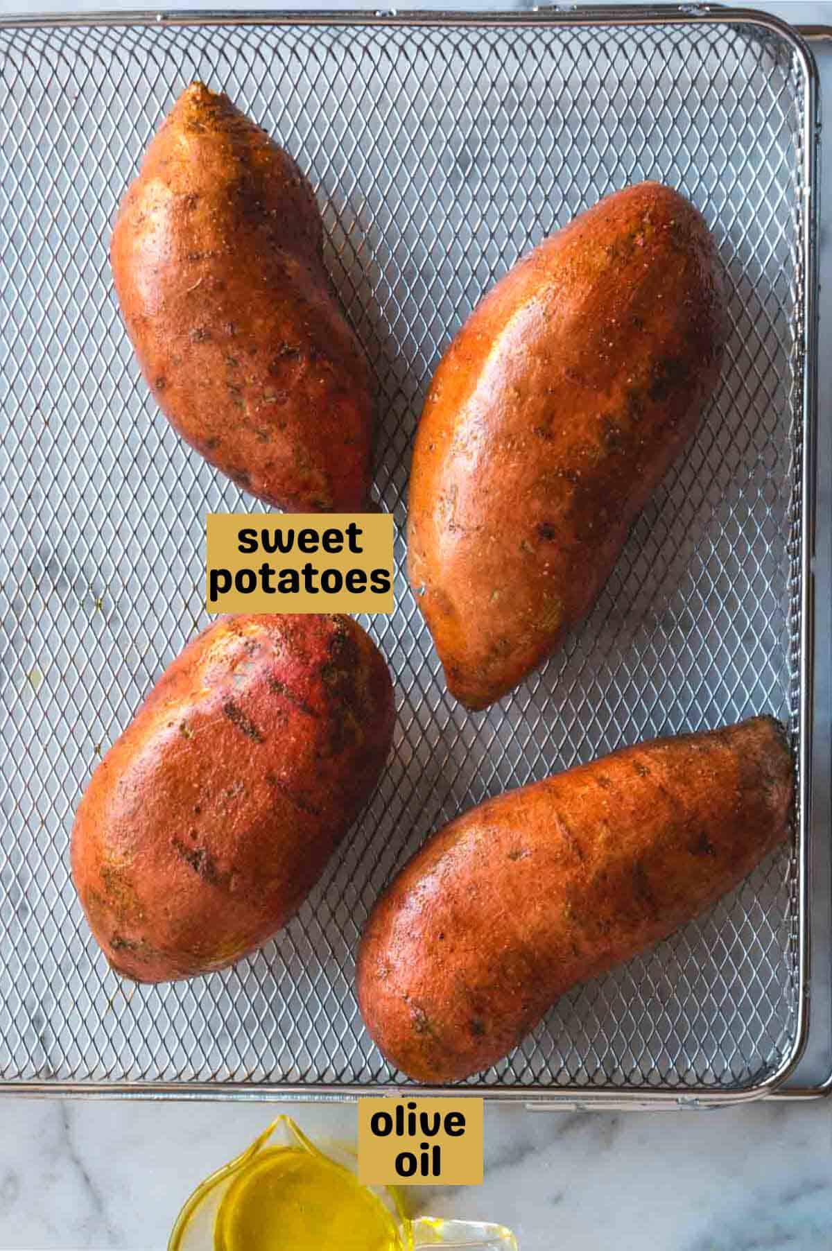 Four whole sweet potatoes on a wire mesh baking sheet next to a small glass cup with olive oil.